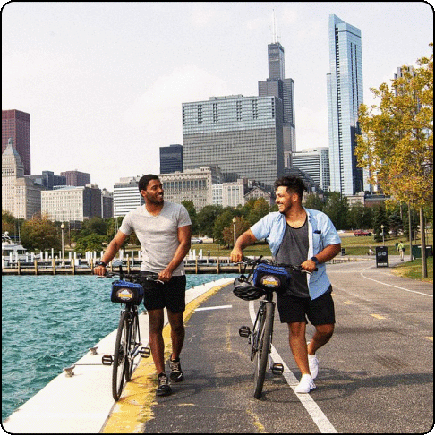 Image Description: Two men smile and look at each other while pushing their bikes on a city bike path along the water.