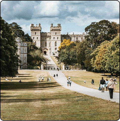 Image Description: Large stone castle with trees and grass interspersed. Groups of people walk along the pathway. 