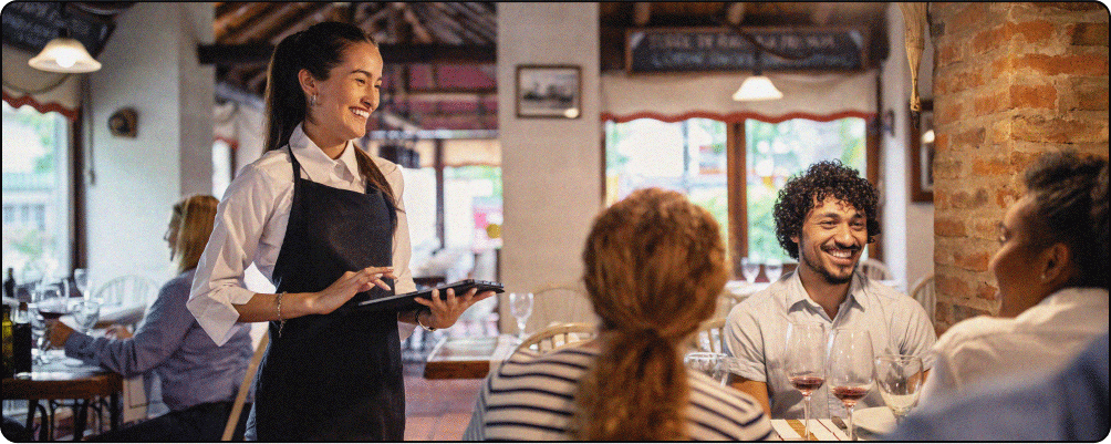 Image Description: A group of 3 people all smile while seated at a restaurant. Their server stands at their table, holding a tablet and smiling at them.