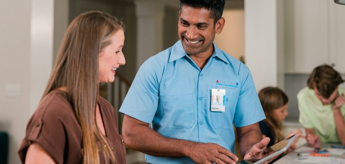 an Aire Serv technician in a blue shirt assisting a woman with long brown hair, while her children do homework in the background