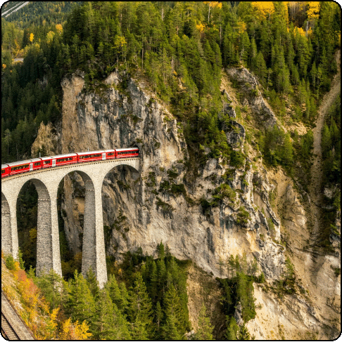 Image Description: A red train cuts through a woodsy gorge and disappears into the side of a mountain.