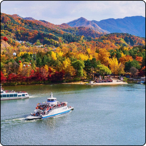 Image Description: There is a calm, large body of water with two boats in the foreground, with mountains and trees in the background showing a wide array of colors from green to orange and yellow.