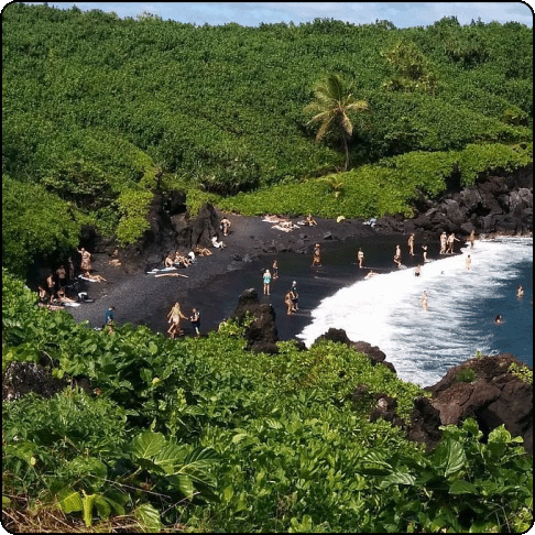 Image Description: A secluded cove with black sands surrounded by lush greenery. People are scattered, laying on the beach and in the water.