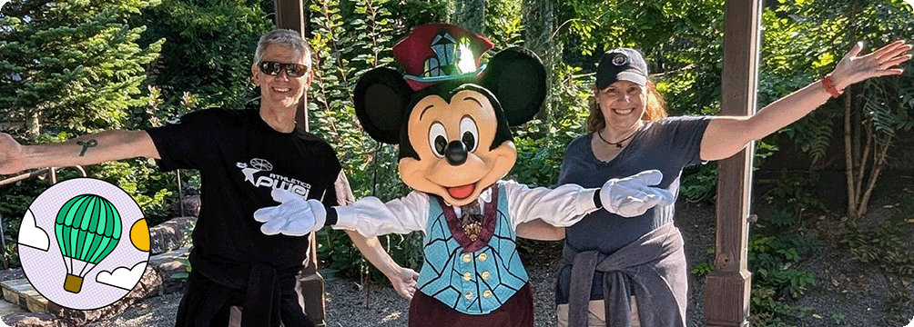 Image Description: Two people posing for a photo with Mickey Mouse. An Experience Explorer badge is on the bottom left corner of the image. 