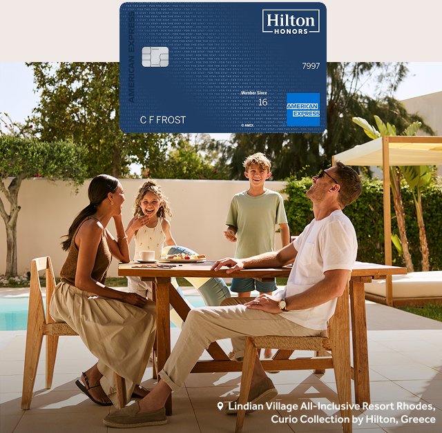 A Hilton Honors Amex Surpass® card sits above a family in laughing at a table by a pool at Lindian Village All-Inclusive Resort Rhodes, Curio Collection by Hilton, Greece.