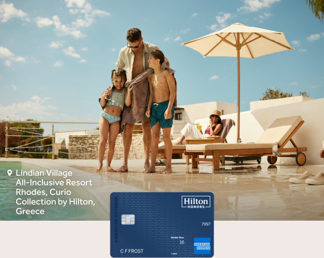 A family stands by a pool at Lindian Village All-Inclusive Resort Rhodes, Curio Collection by Hilton, Greece with a Hilton Honors Amex card visible.