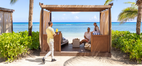 Two people relax in a beachside cabana while another stands outside with drinks, with the ocean in the background.