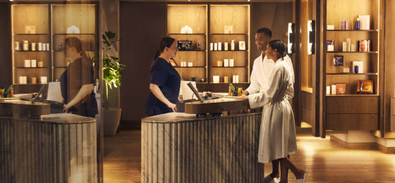Two people in white robes stand at a marble counter in a reception area, with a staff member behind the counter.