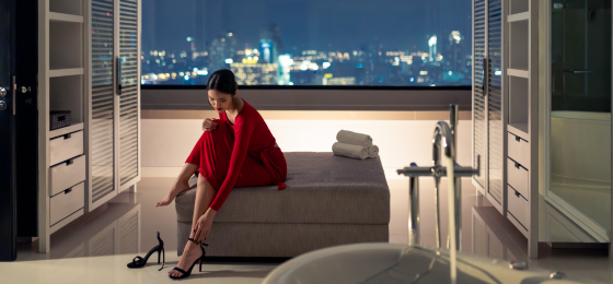 A person in a red dress sits on a bed in a modern room at night, putting on black high heels.
