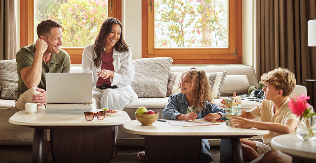 A family spends time in a living room; adults sit on a couch while children draw and play at a table.