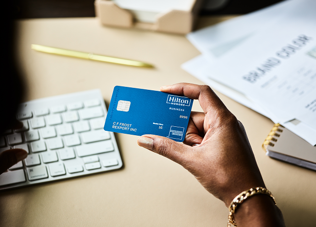 A person holds a blue Amex Hilton Honors Business Card at a desk.