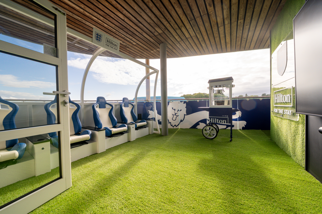 Outdoor cinema area with an England football team theme, featuring seating under a wooden canopy, artificial grass, and a Hilton branded popcorn machine.