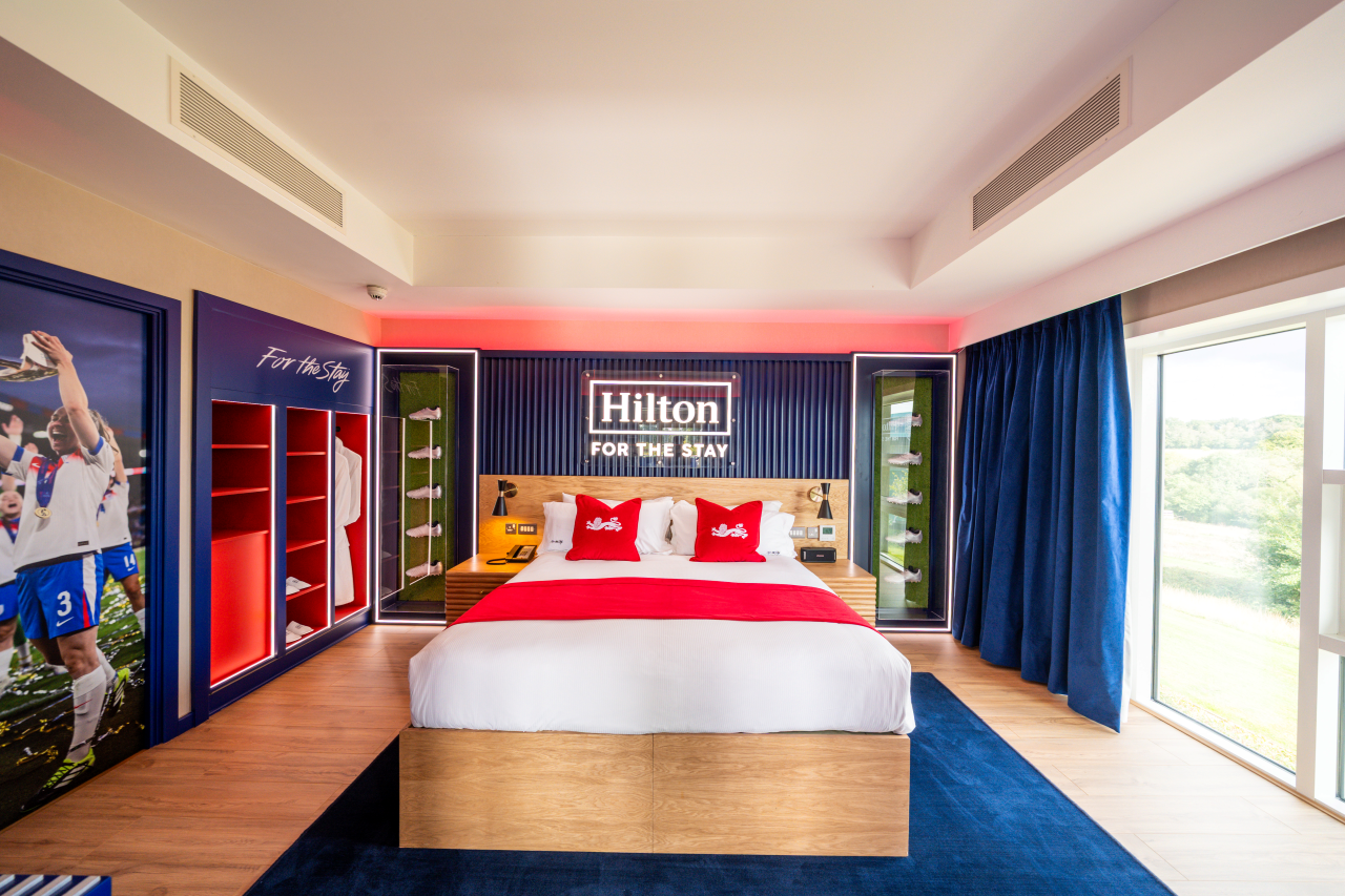 Hotel room with a large bed featuring white sheets and red pillows featuring lion logo of the England football team.
