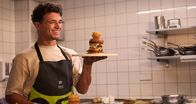 A person in a kitchen holding a gourmet burger on a plate.