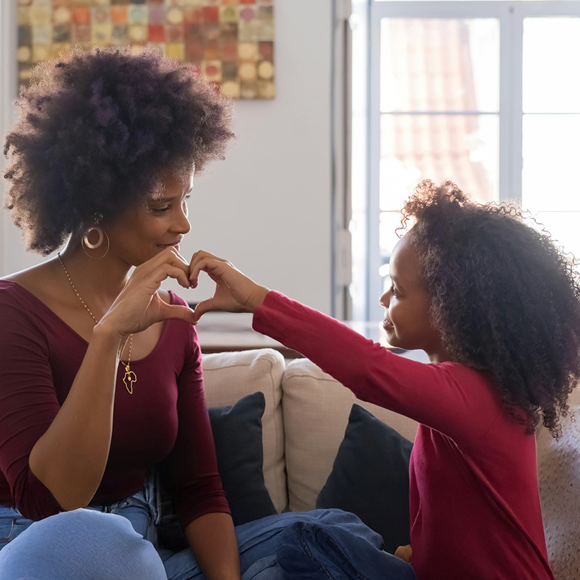 A mother and young daughter sit together on a sofa, joining their hands to form a heart shape symbolizing American Heart Month.
