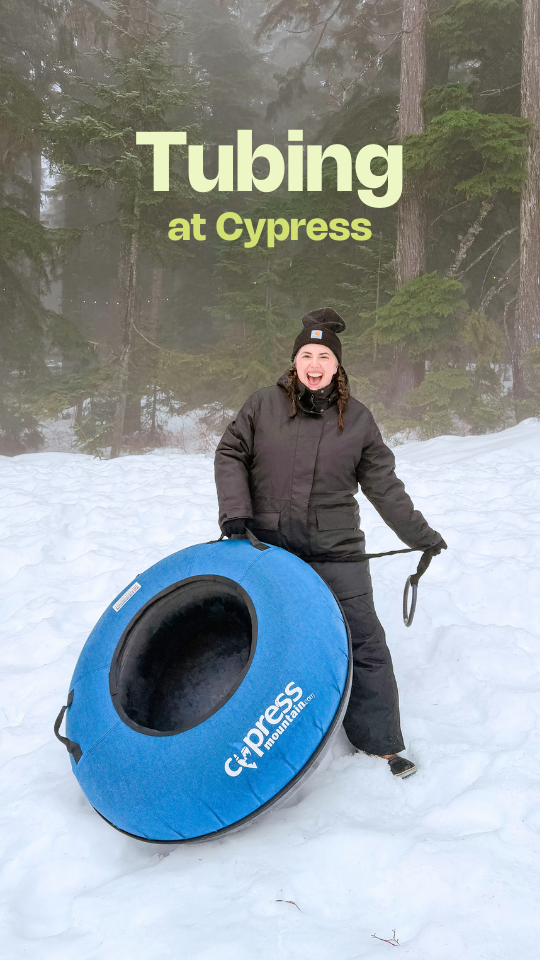 A smiling woman stands in the snow at Cypress Mountain, holding a blue snow tube with forest trees in the background and the text 