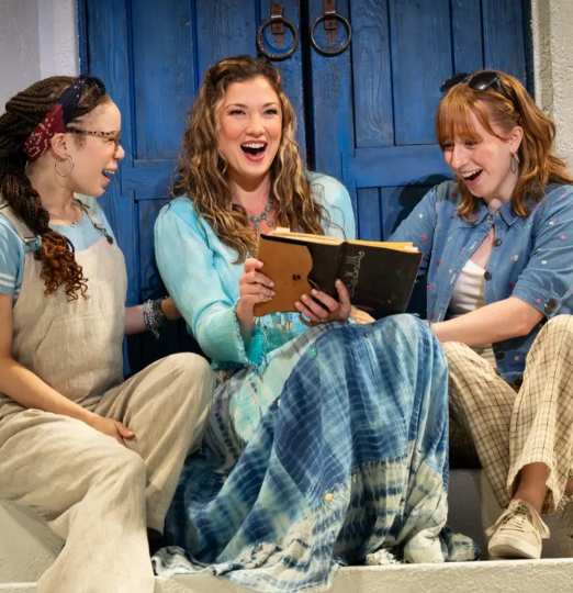 Three women sit on steps in front of bright blue doors, laughing together as they look at an open book one of them is holding.