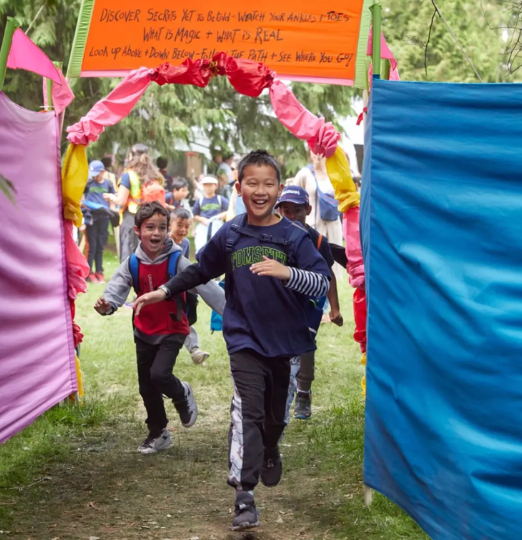 Children run joyfully through a colorful outdoor entrance at the Vancouver International Children's Festival, with bright fabric panels and a hand-painted sign overhead.