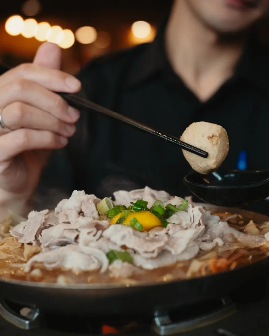 A person uses chopsticks to lift a meatball from a steaming hot pot filled with sliced meat, egg yolk, and green onions in a warmly lit restaurant.