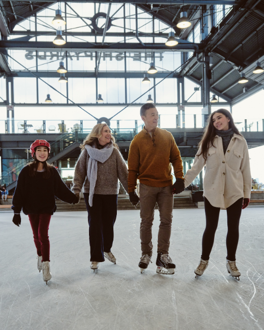 A family of four ice skates hand in hand at The Shipyards in North Vancouver, inside a covered outdoor rink with industrial beams and hanging lights overhead.