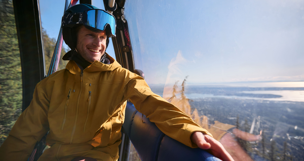 A smiling man in a yellow ski jacket and helmet with goggles sits inside a gondola, looking out at a sunny mountain and city view below.