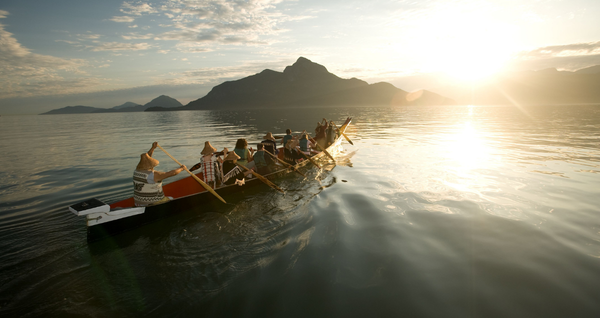 People paddling a traditional canoe across calm coastal waters at sunset, with mountains silhouetted in the background.