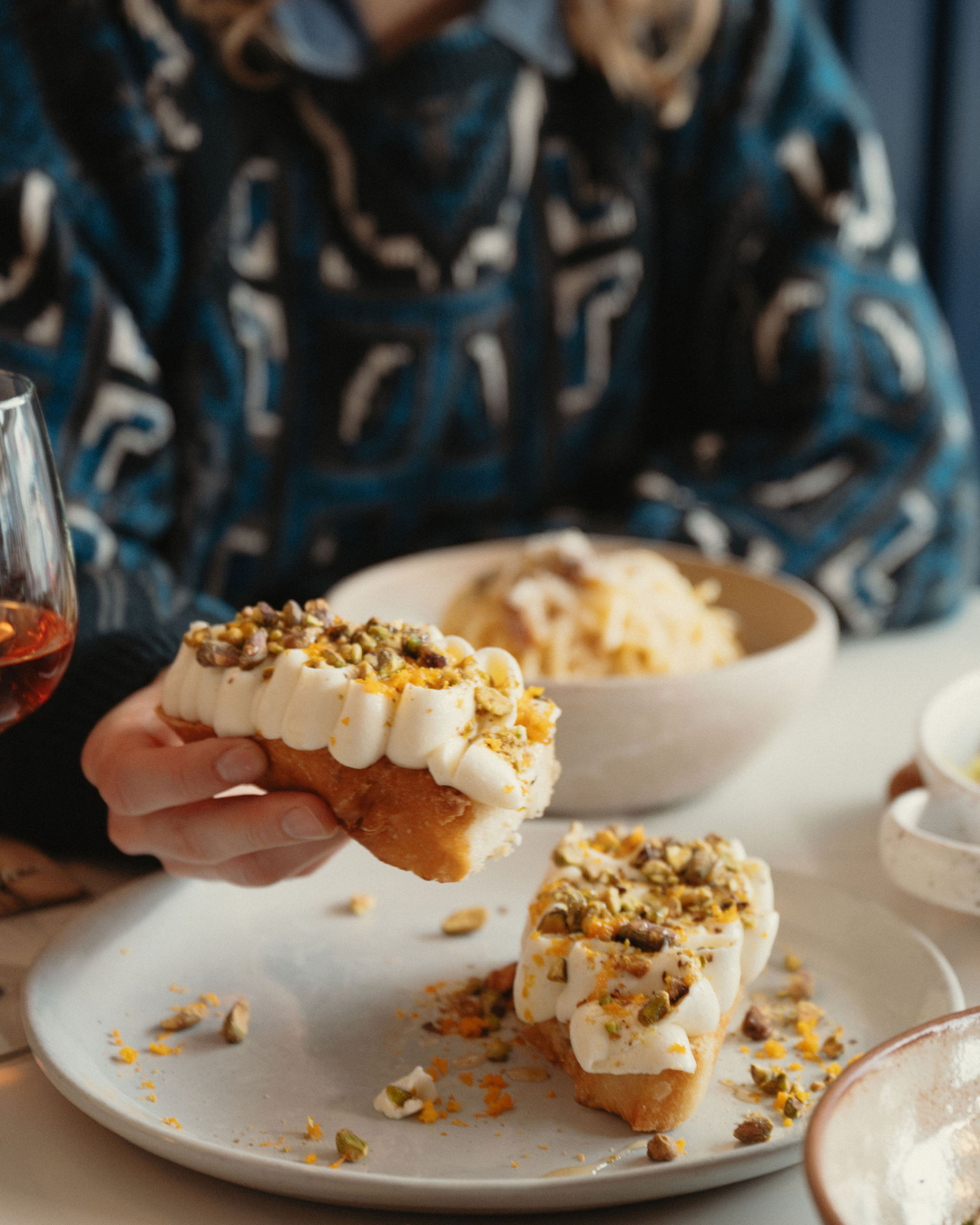 Close-up of a hand holding a pastry topped with whipped cream, pistachios, and citrus zest, with a second pastry and a glass of wine on the table.