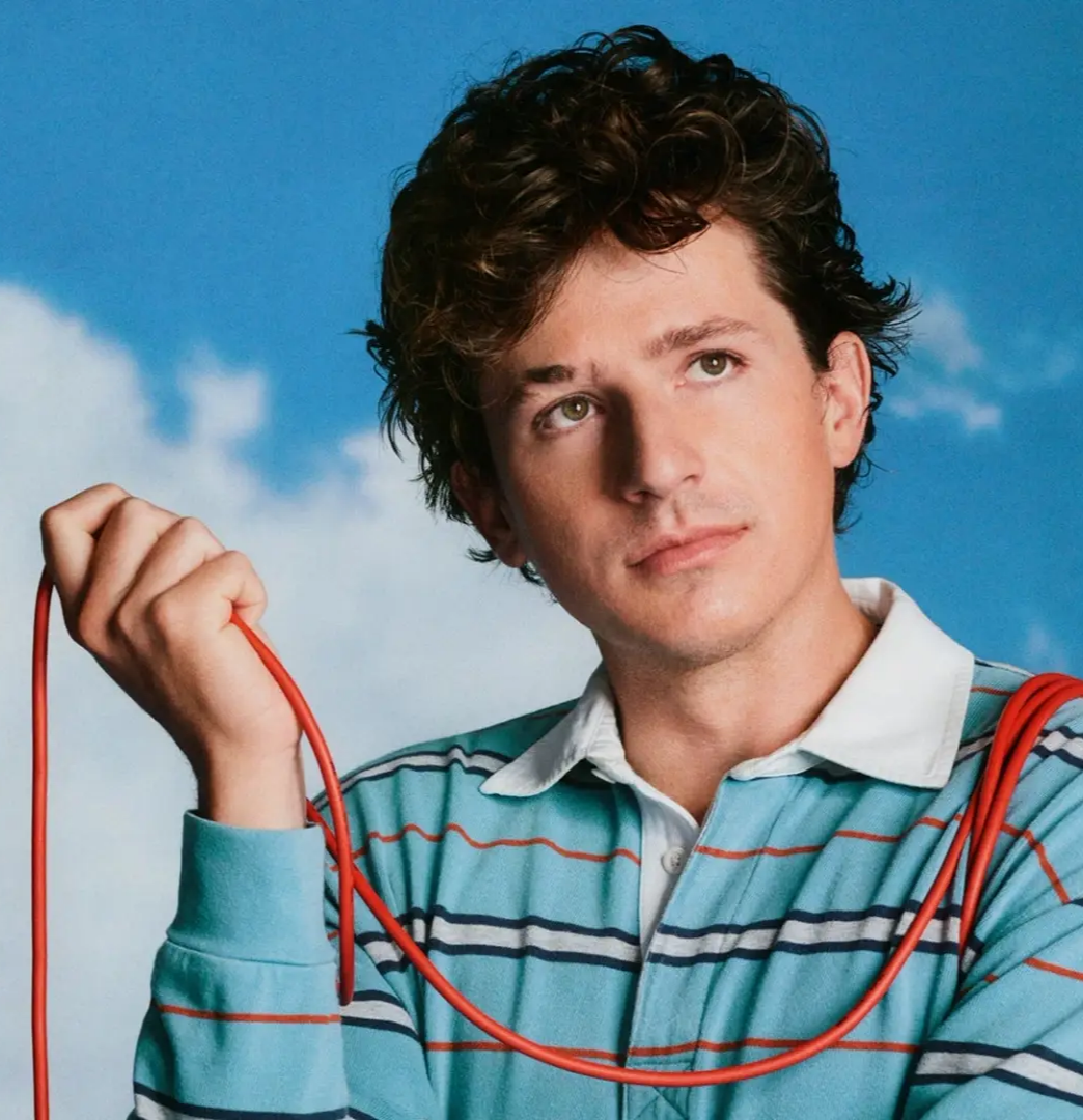 Portrait of Charlie Puth with curly hair holding a red cable, wearing a striped polo shirt, posed against a bright blue sky background.