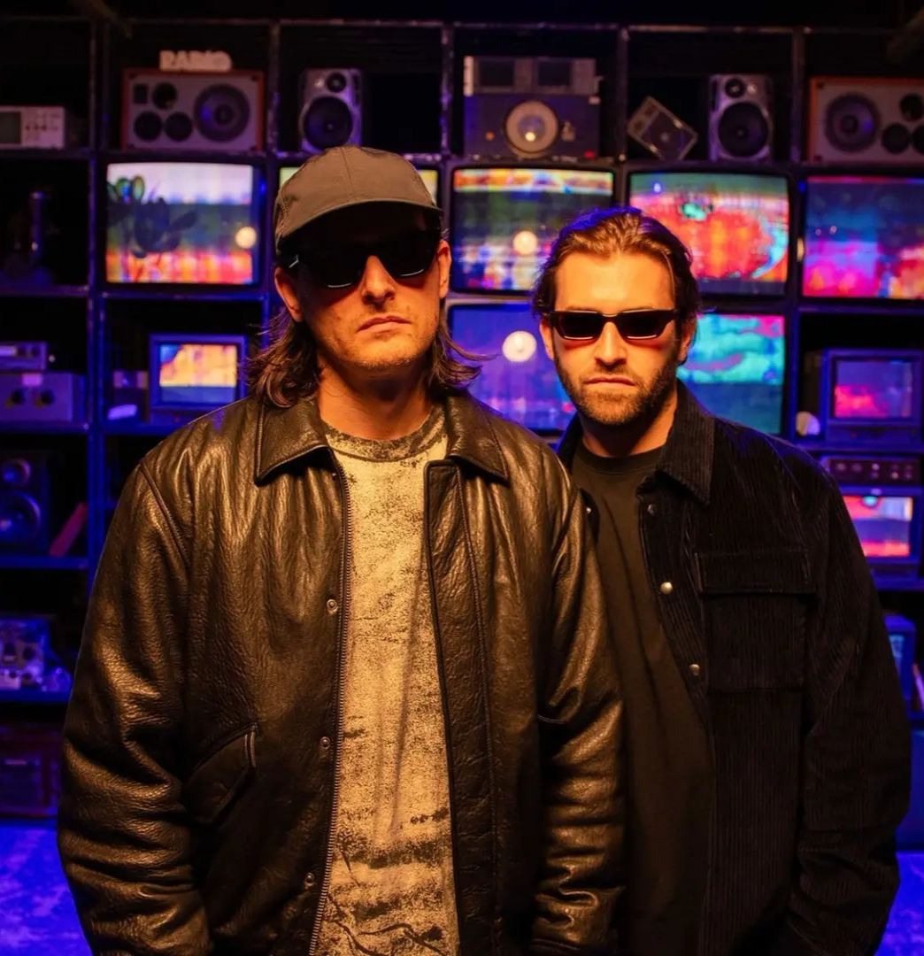 Promotional image of Zeds Dead featuring two artists wearing sunglasses and dark clothing, standing in front of a wall of colorful screens and speakers.