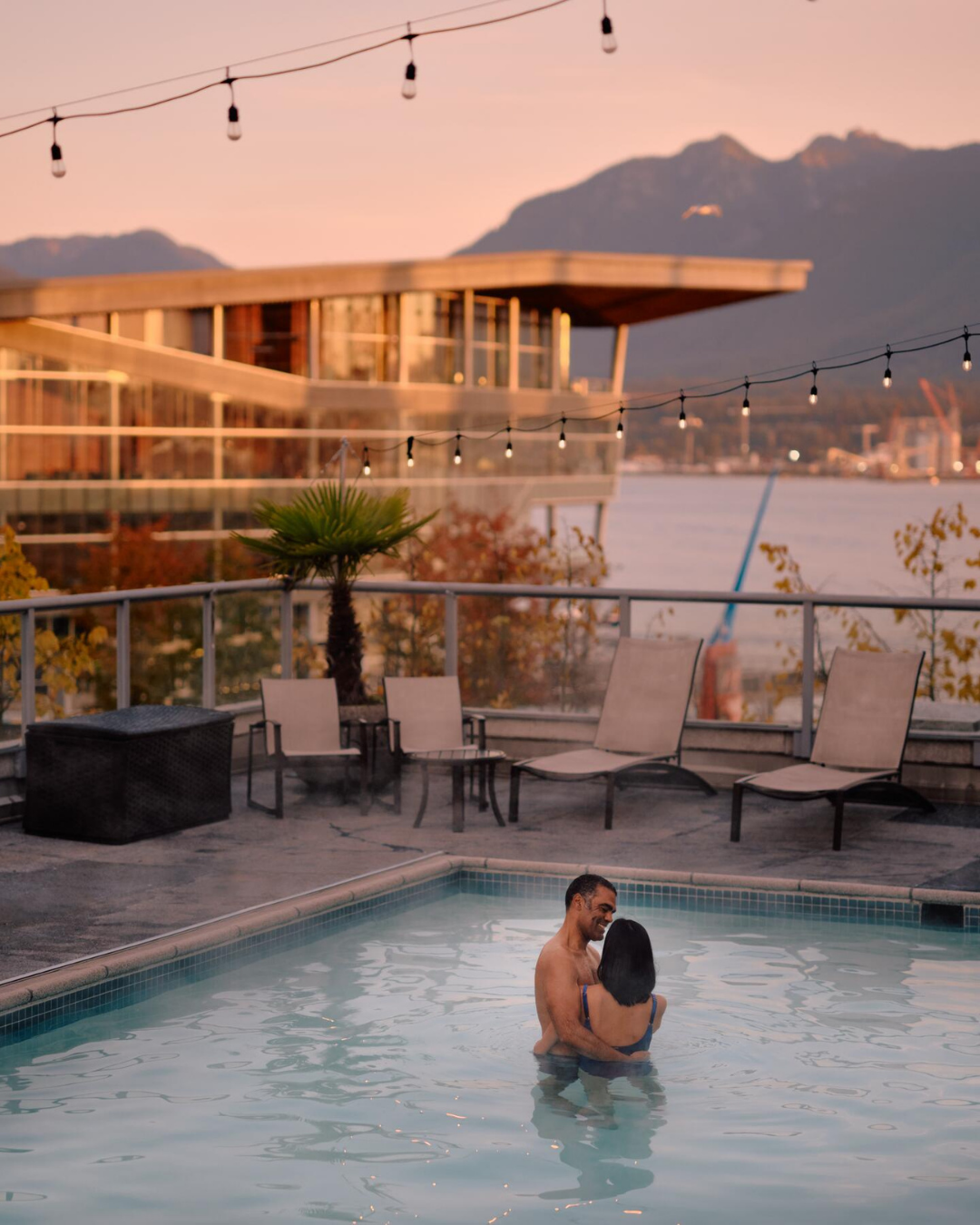 Couple embracing in a heated rooftop pool at sunset in Vancouver, with string lights, mountains, and the waterfront behind them.
