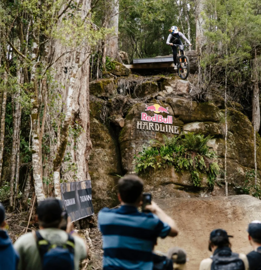 A downhill mountain biker launches off a cliffside drop at Red Bull Hardline in British Columbia, as a crowd watches from below.