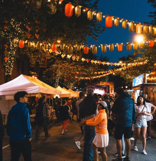 People explore a lively evening street festival in Vancouver’s Chinatown, with glowing lanterns and vendor tents lining the street.