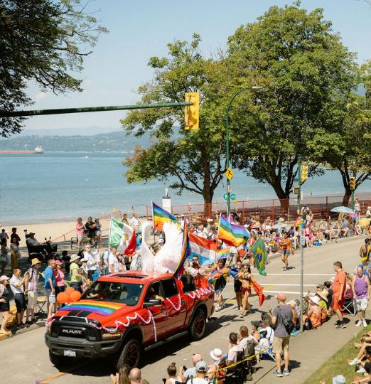 A red truck decorated with rainbow flags leads a Pride parade along Vancouver’s waterfront, with people waving flags and crowds gathered along the street.