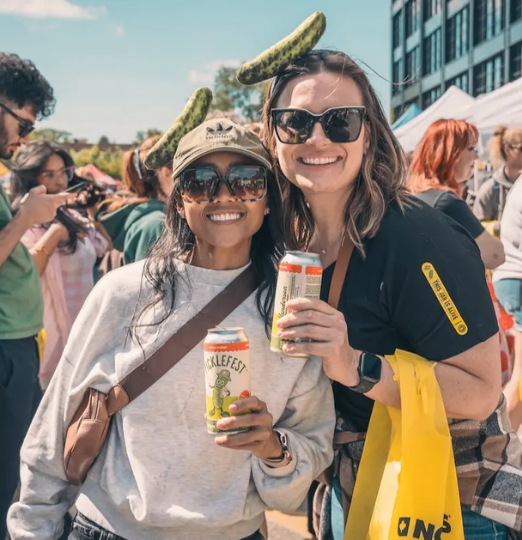 Two women pose with canned drinks at a sunny outdoor festival in Vancouver, wearing sunglasses and playful pickle headbands.