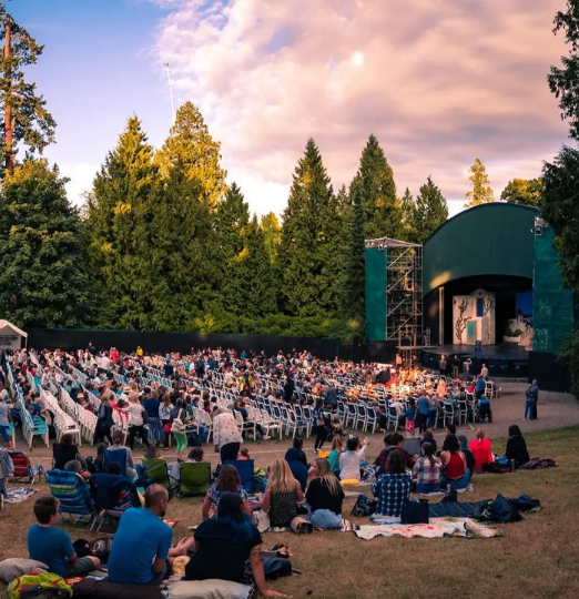 An outdoor theatre performance in Stanley Park with a large audience seated on chairs and blankets at sunset.