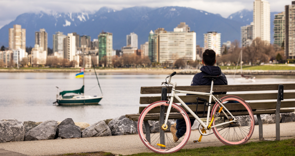 Person relaxing on a waterfront bench beside a bicycle, overlooking Vancouver’s skyline, marina, and North Shore Mountains.