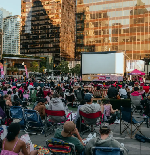 Crowd gathered in downtown Vancouver for an outdoor summer movie screening surrounded by city skyscrapers at sunset.