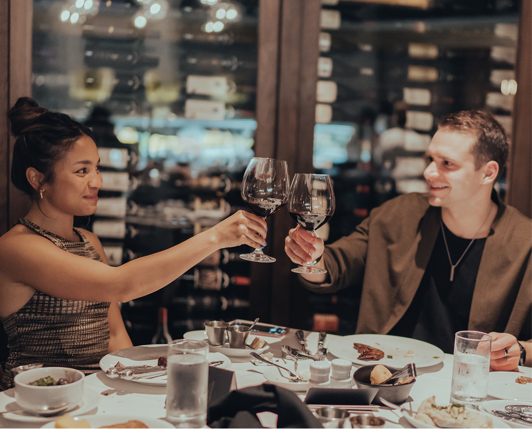 two people holding up wine glasses to toast over a table set with food 