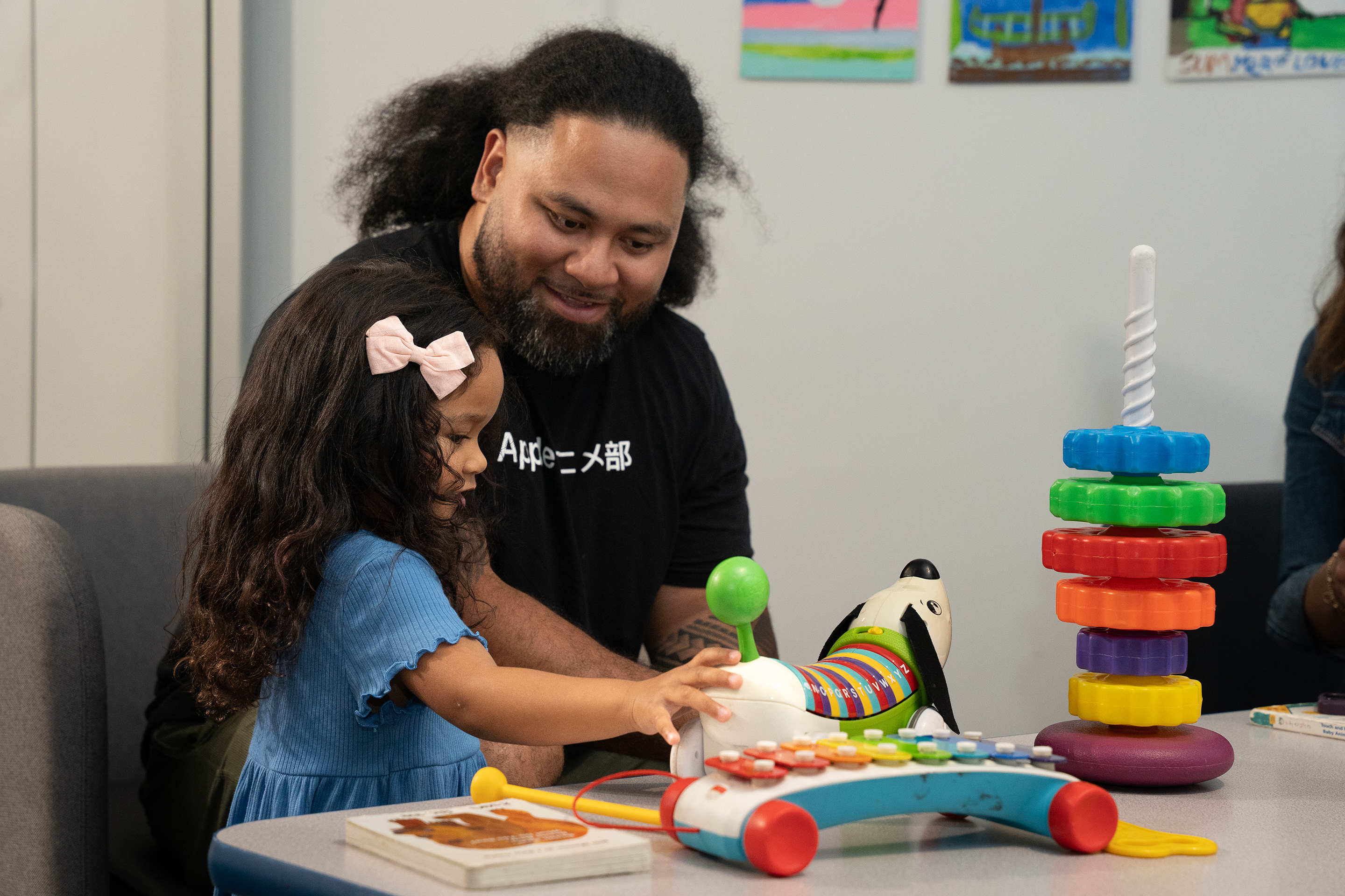 Adult sits beside a young child playing with colorful educational toys at a table, including a stacking ring toy, xylophone, and interactive activity toy, in a bright playroom.