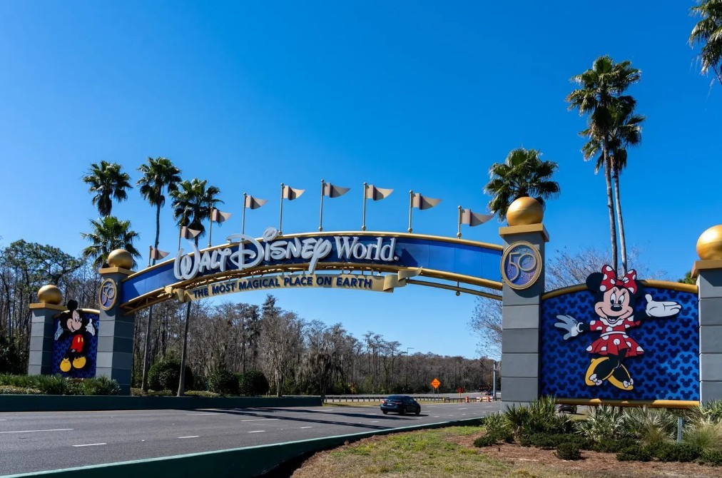 Entrance arch to Walt Disney World featuring Mickey Mouse and Minnie Mouse, with palm trees, blue skies, and a multi-lane roadway leading into the resort.