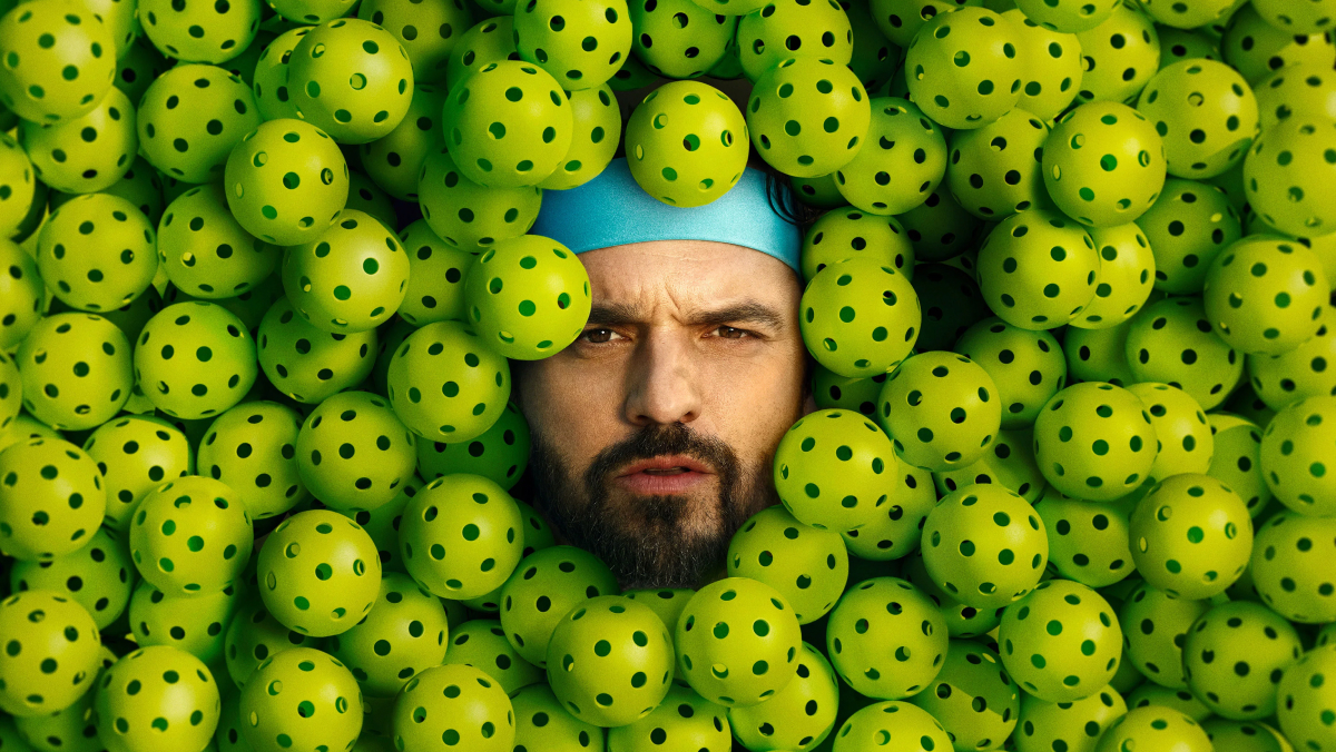 This is a promotional image for the 2026 pickleball-themed comedy movie The Dink. It features actor Jake Johnson looking up at the camera with exasperation. Only his face is visible because he's buried in pickleballs.