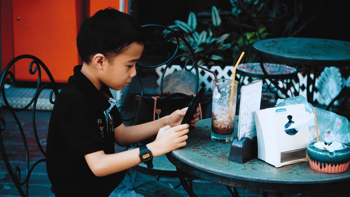 This is a photo of an elementary school boy sitting at a table. He's looking down at a handheld digital device.