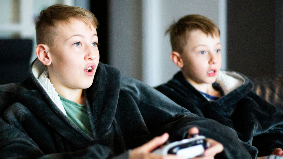 This is a photo of two adolescent boys playing video games. They're holding controllers and looking toward a screen with excited expressions on their faces.