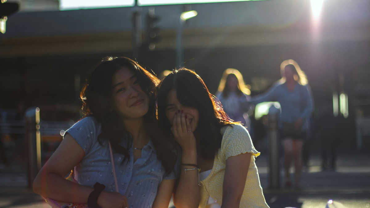 This is a photo of two adolescent girls. One smiles and looks at the camera while the other laughs with her hand in front of her face.