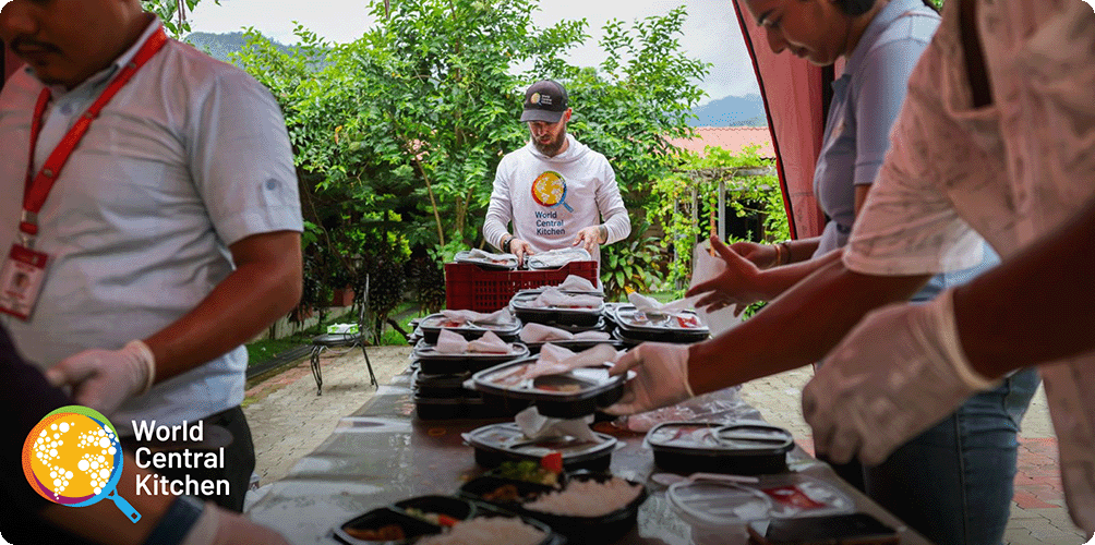 Image Description: World Central Kitchen crew members packing and preparing to-go meals at a long table with greenery in the background. World Central Kitchen logo in the bottom left corner of the image.